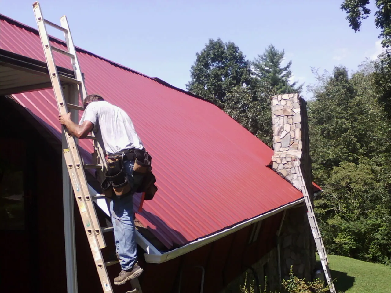 Roofer on a ladder beside a red metal roof and stone chimney — Soffit & Fascia Repair by Roofing Brothers in Charlo